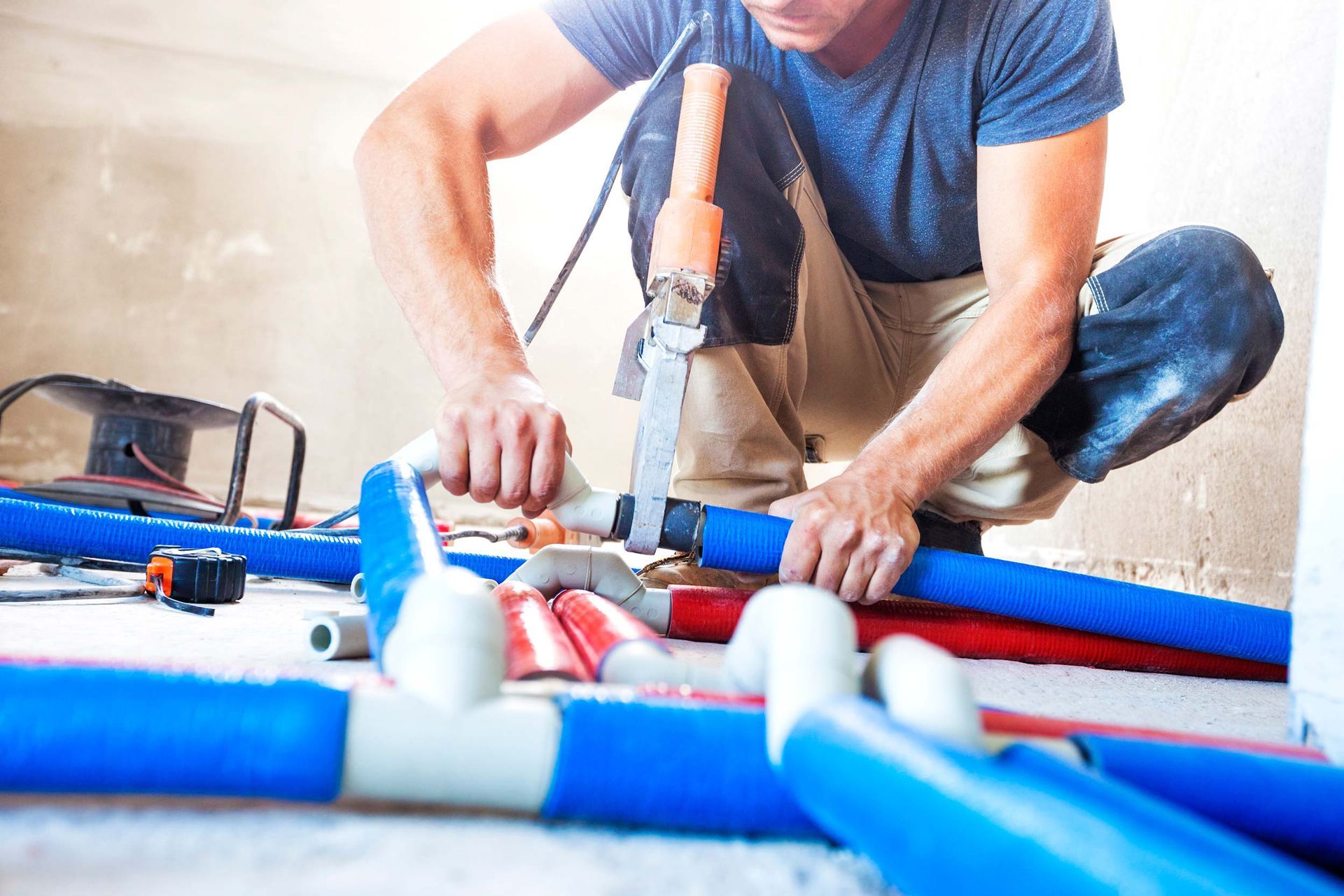 Plumber kneeling, connecting blue pipes with a tool on a construction site.