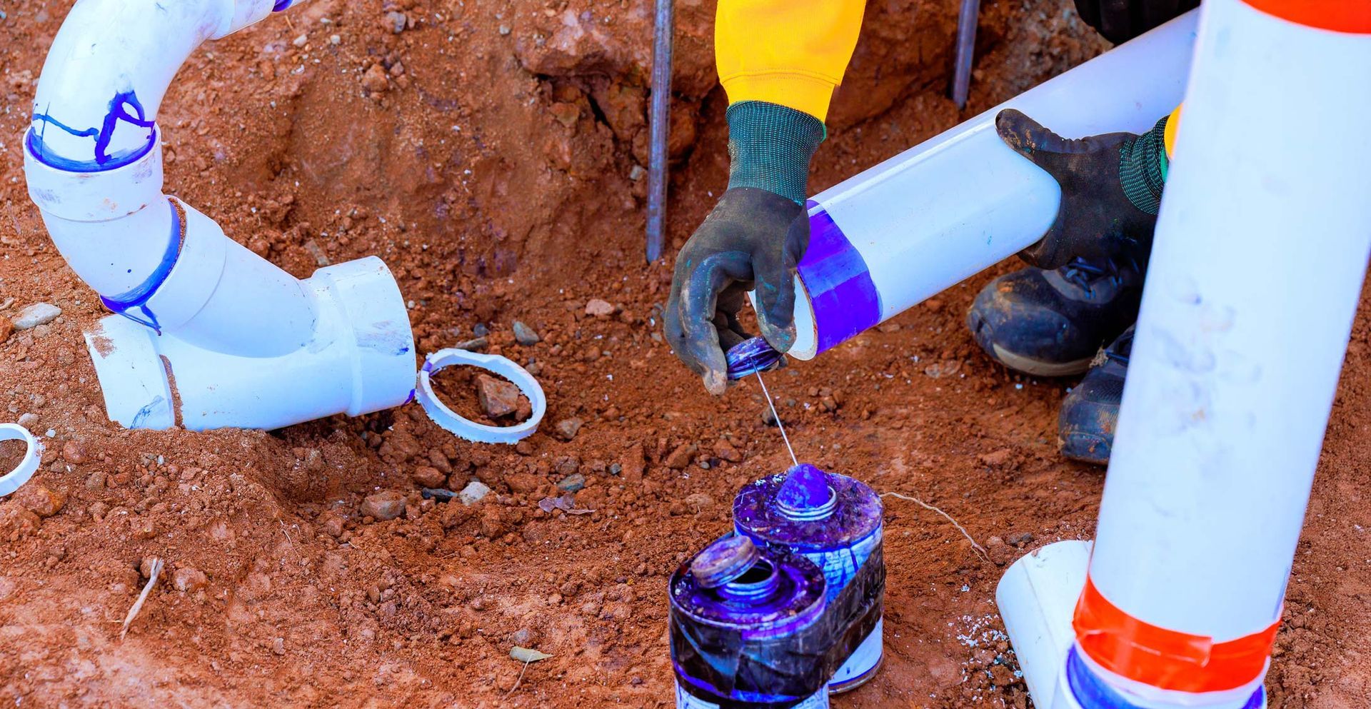 Plumber gluing PVC pipes in a dirt trench, applying purple primer.