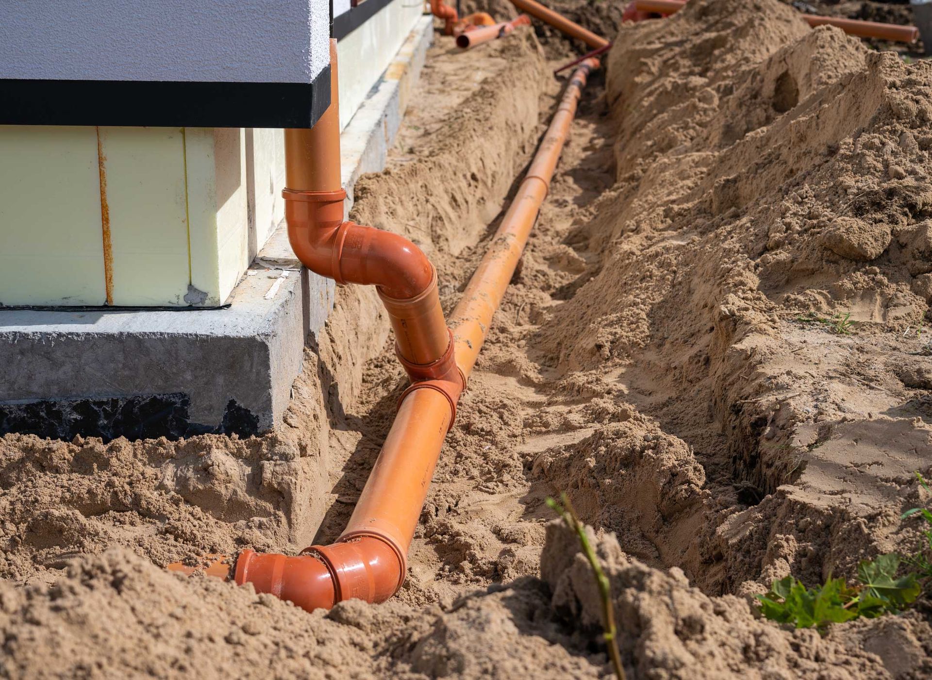 Orange drainage pipes in a trench next to a building, partially buried in sand.