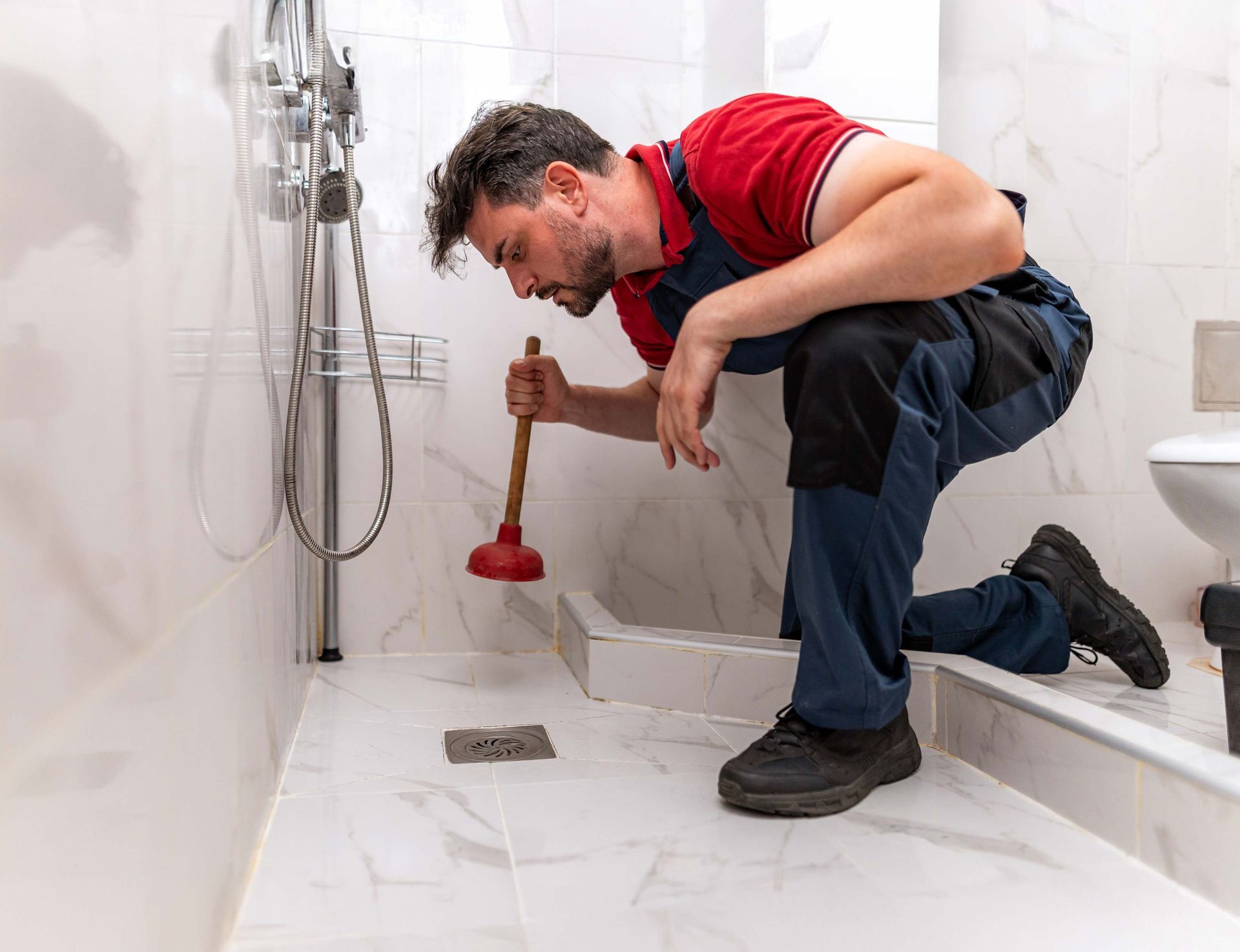 Plumber using a plunger in a white-tiled shower, inspecting the drain.