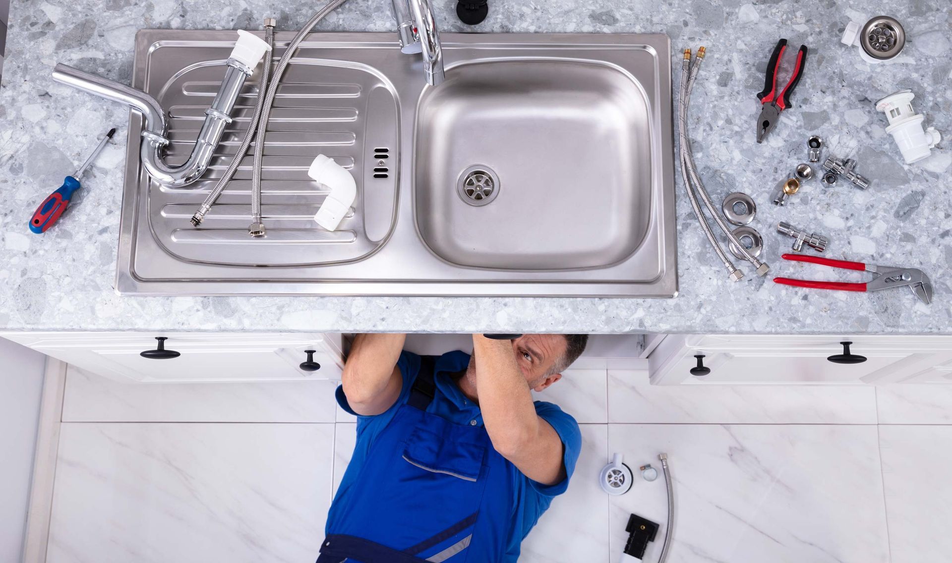 Plumber working under a kitchen sink with tools. Sink is stainless steel, countertop is speckled, person in blue overalls.