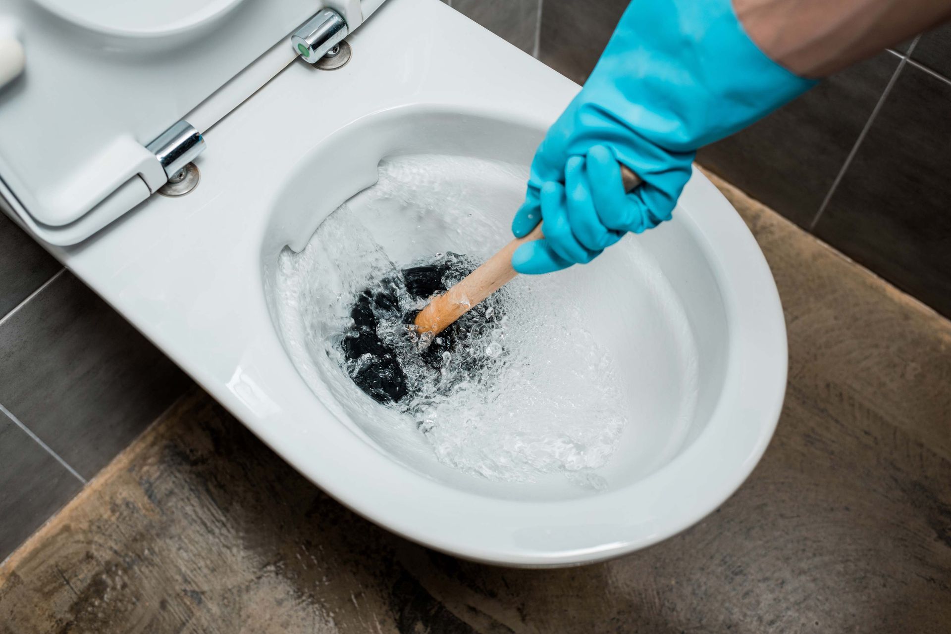 Person in blue glove plunges a clogged toilet, white bowl, wood and tile background.
