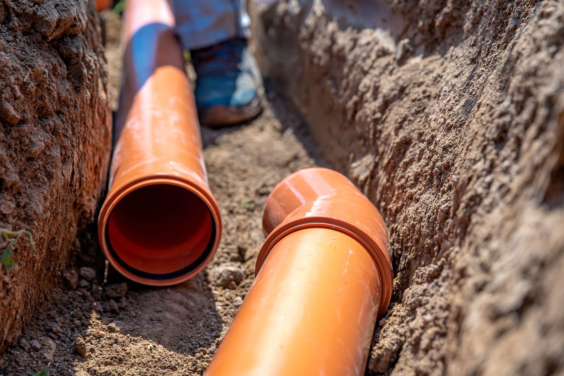 Orange pipes in a trench, prepared for plumbing, with a person's legs and shoes visible.