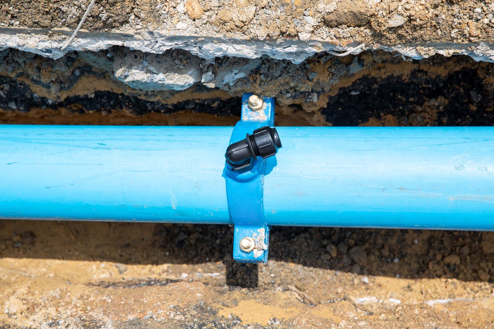Blue water pipe with black fitting and supporting clamp in a trench.