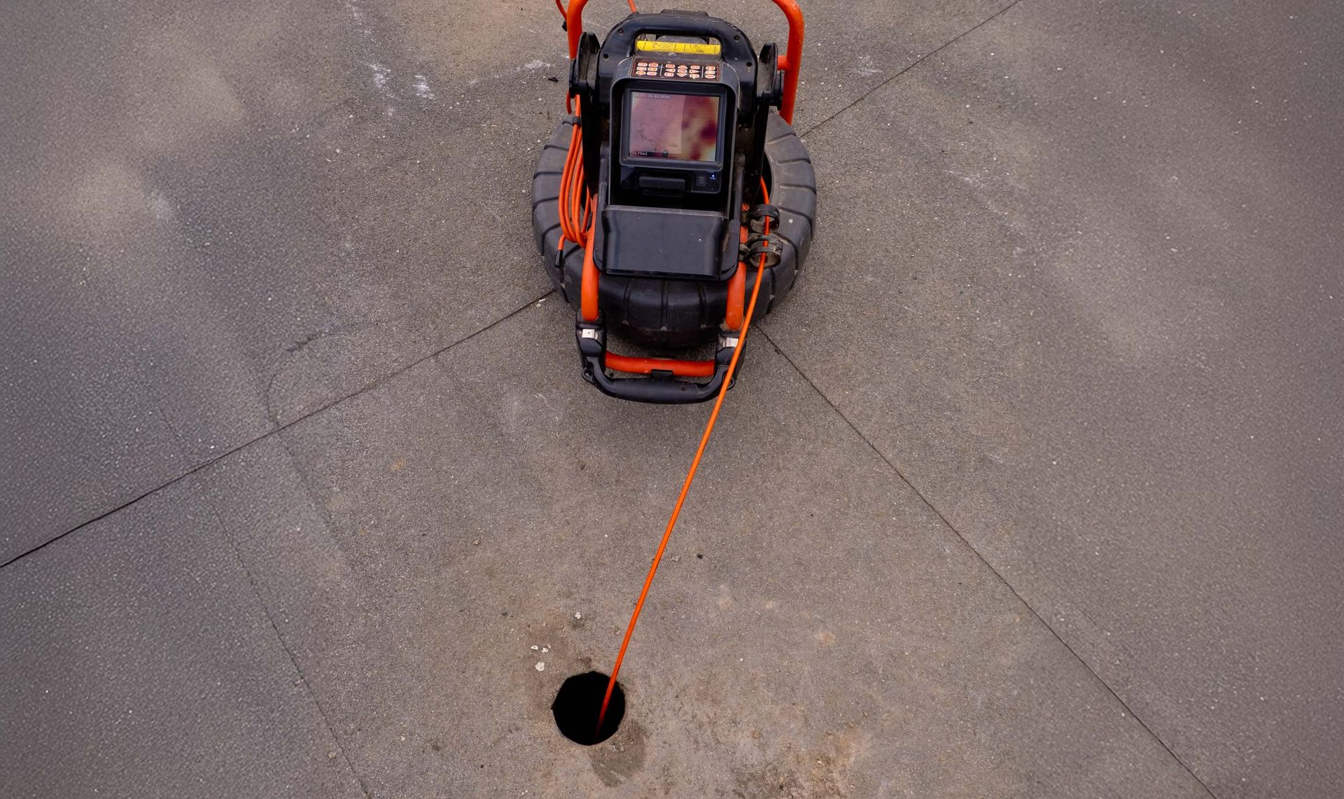 Overhead view of an orange and black machine drilling into asphalt, with a safety line extending from the machine.