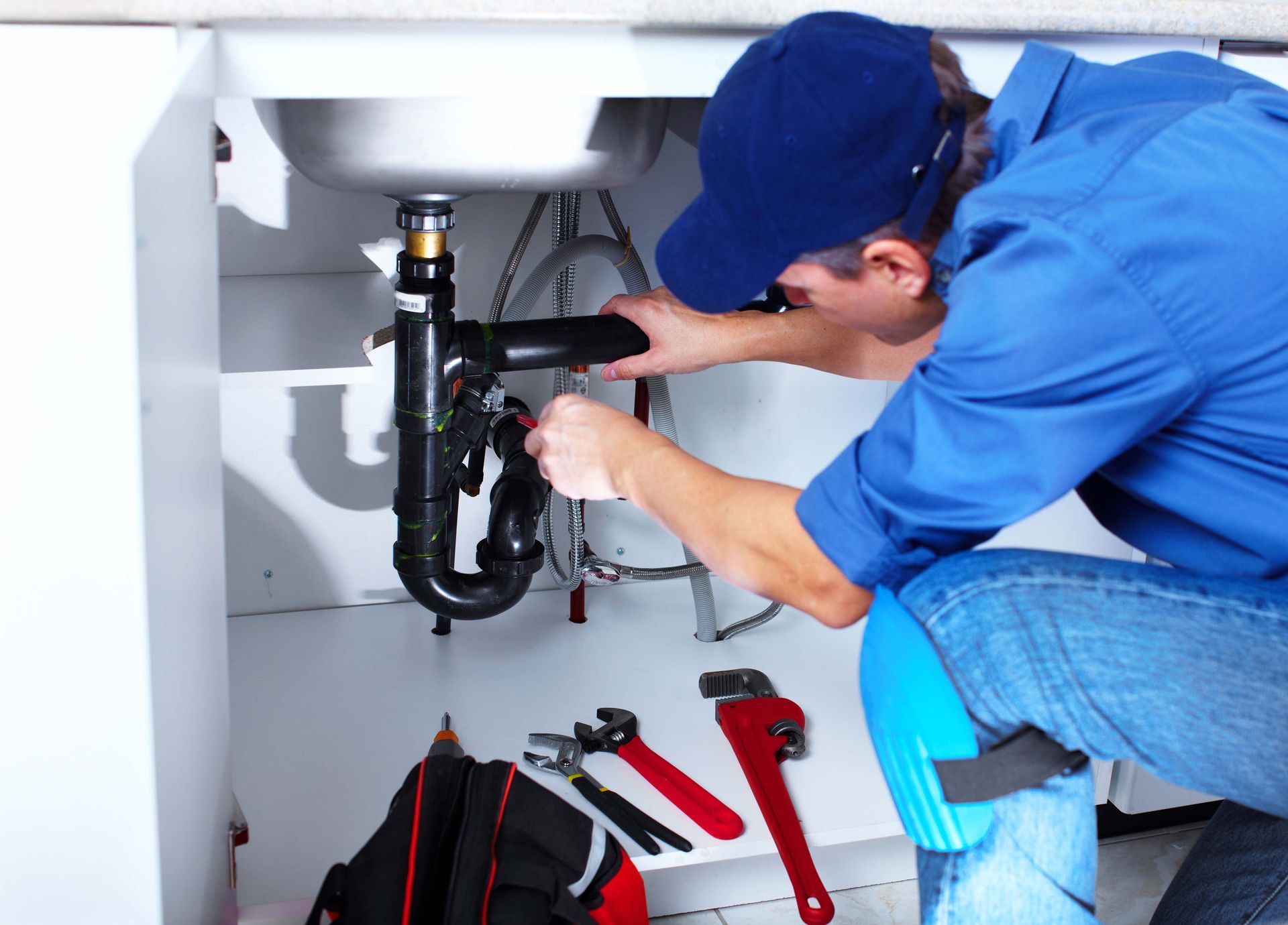 Plumber in blue shirt and cap working under a sink, using tools.