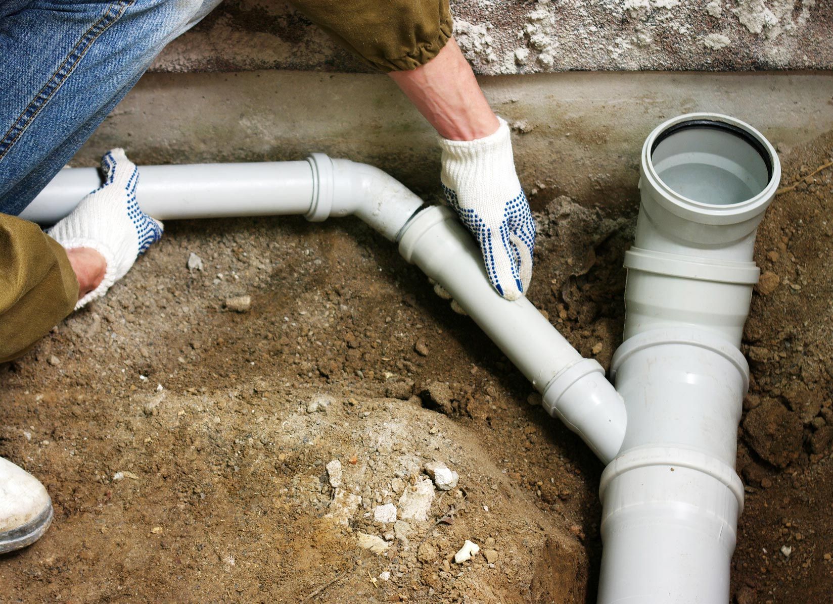 Person installing gray PVC pipes in a dirt trench, wearing gloves.