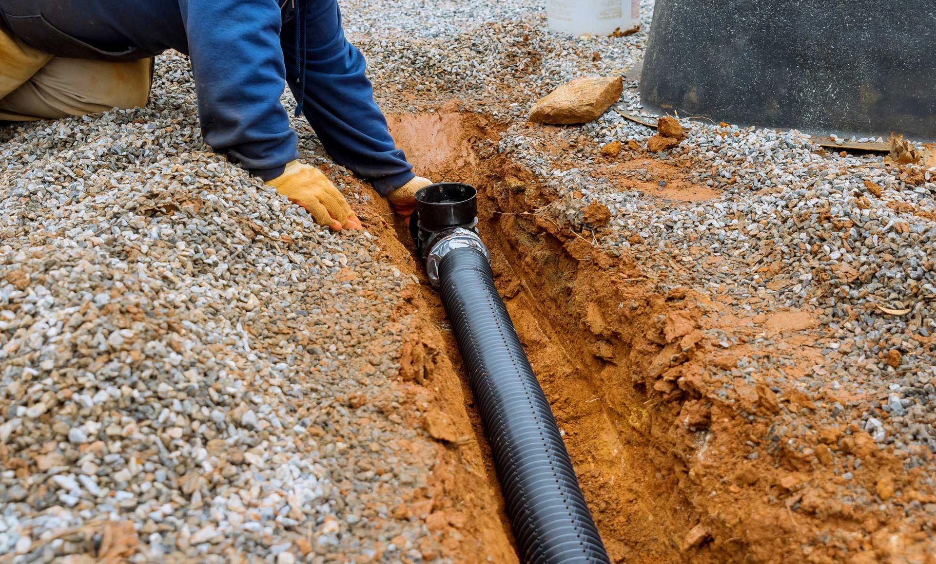 Person installing corrugated black drain pipe in a trench, surrounded by gravel and soil.