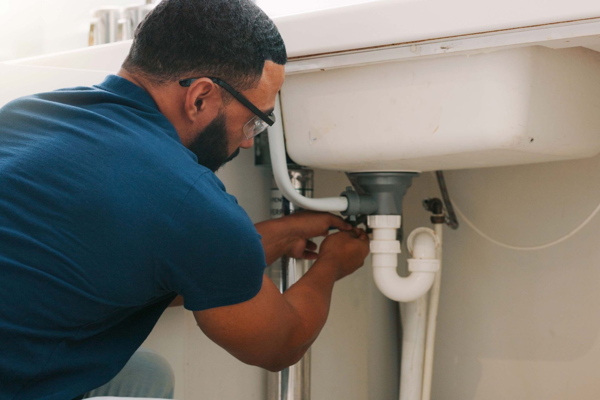 A person in blue shirt works on the plumbing under a white sink, tightening a pipe.