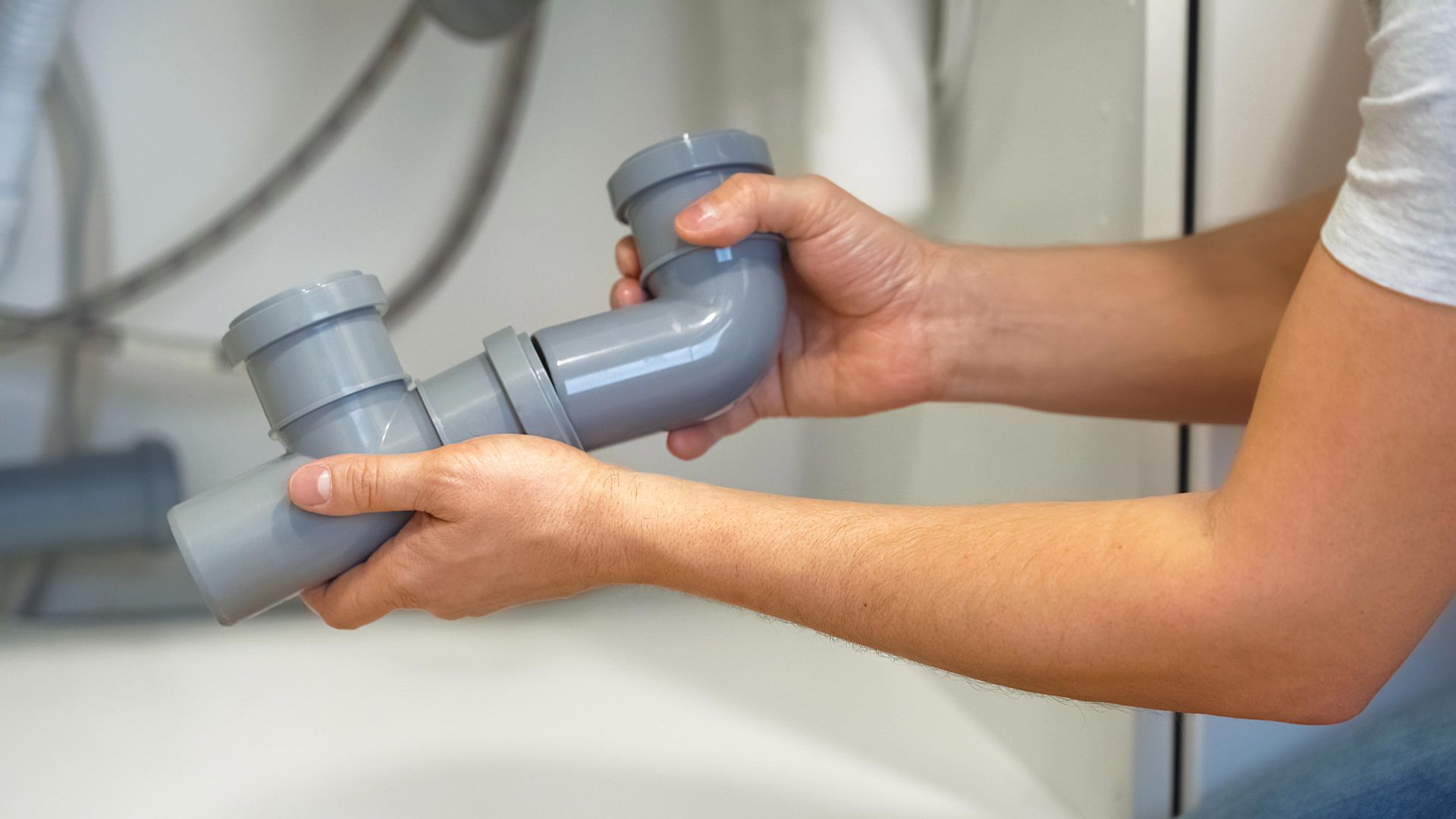 Person holding gray PVC plumbing trap with curved sections, likely for a sink drain, against a white background.