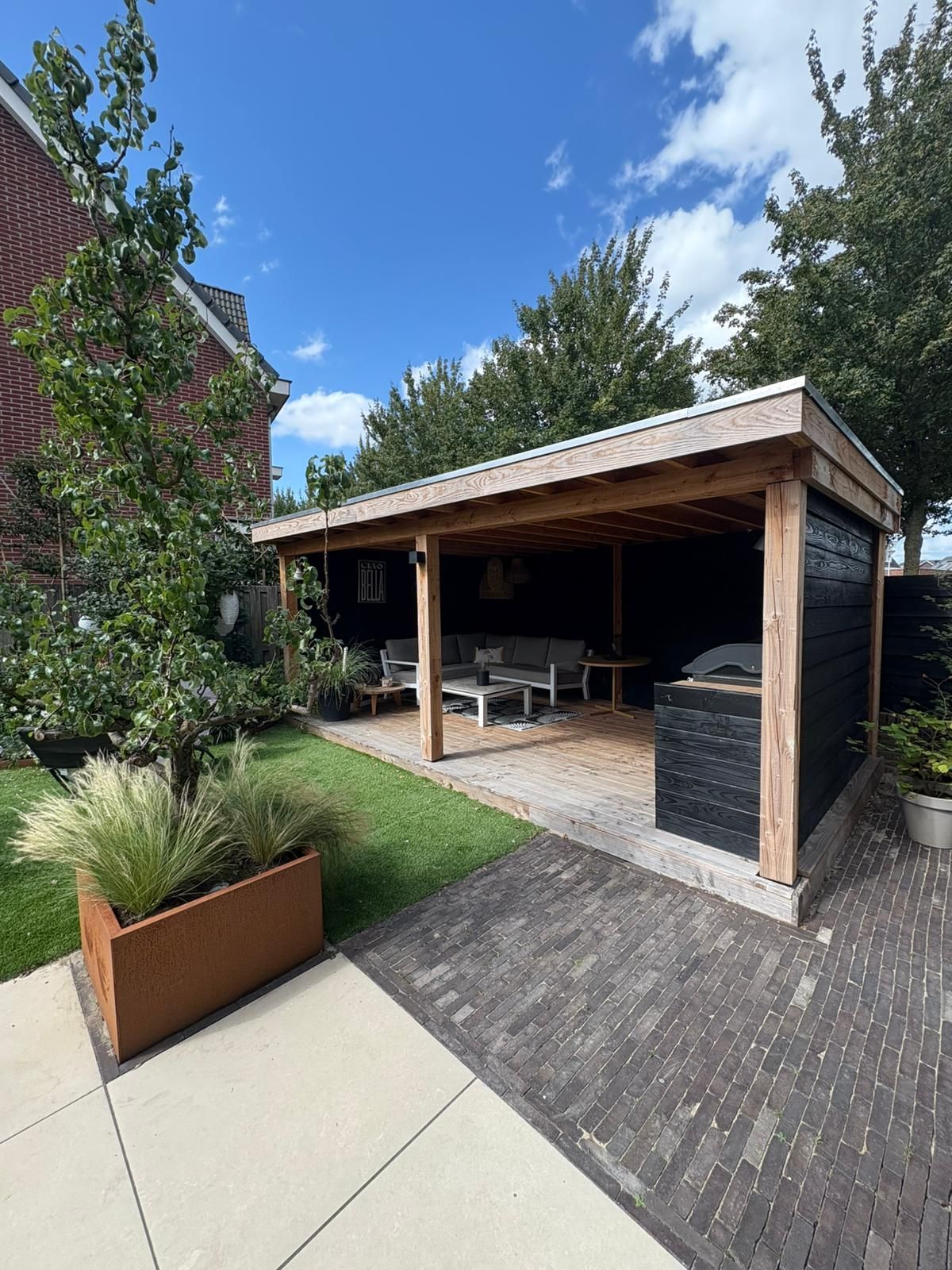 Een terras in de buitenlucht met zitgedeelte, barbecue en een stenen pad onder een houten overkapping op een zonnige dag.