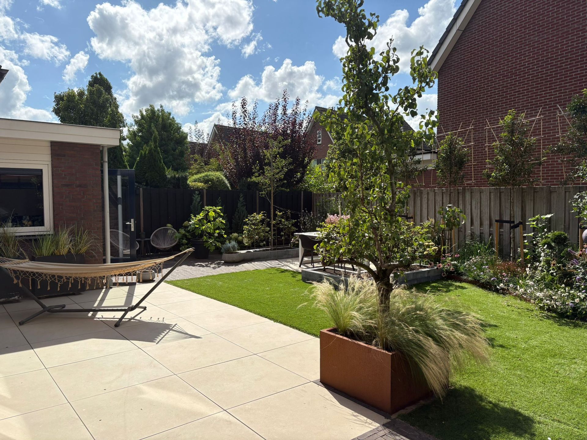 Terras in de achtertuin met gazon, bomen en een hangmat op een zonnige dag.