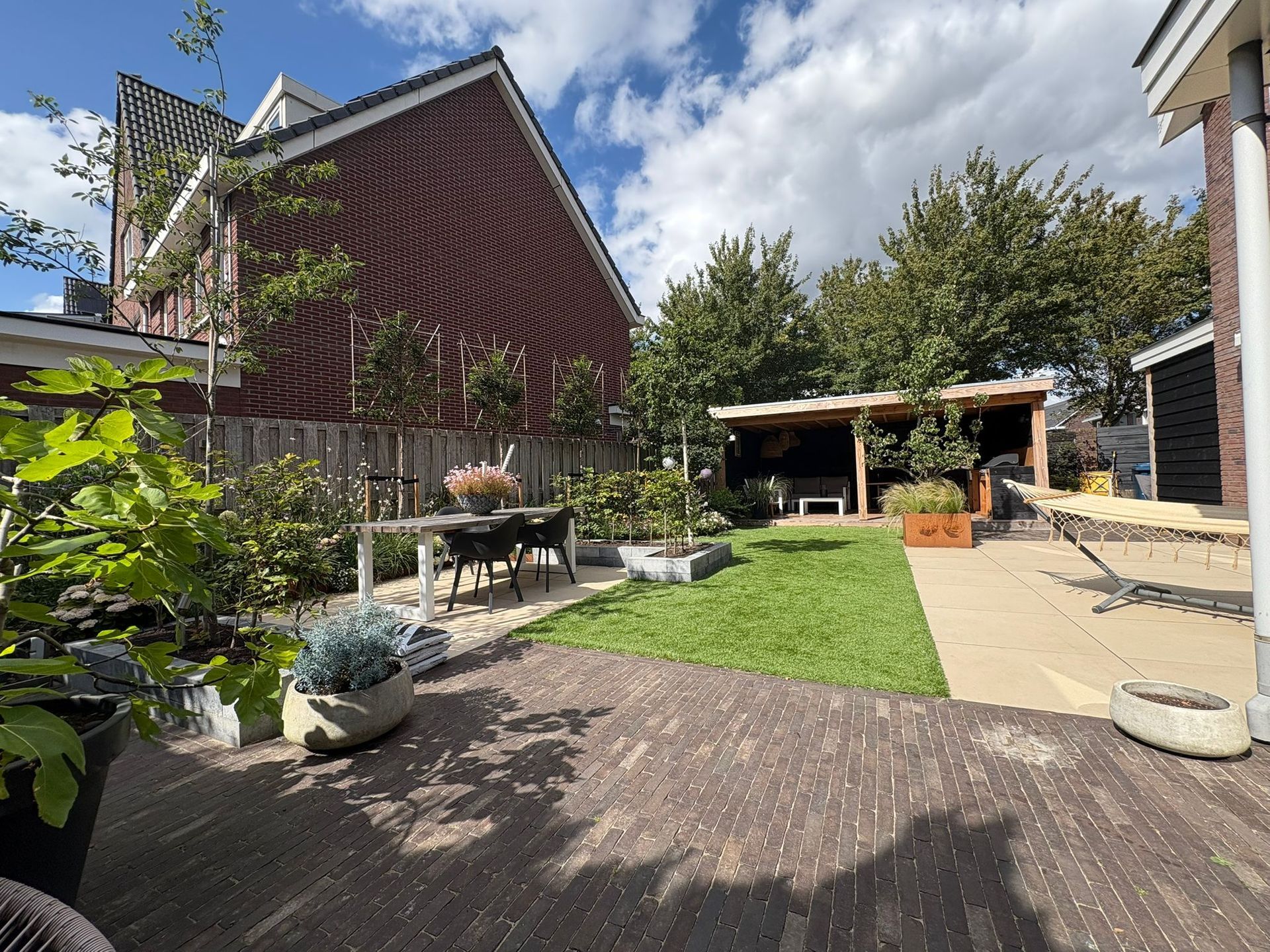 Achtertuin met stenen terras, klein gazon, houten schutting en schuur, onder een gedeeltelijk bewolkte hemel.