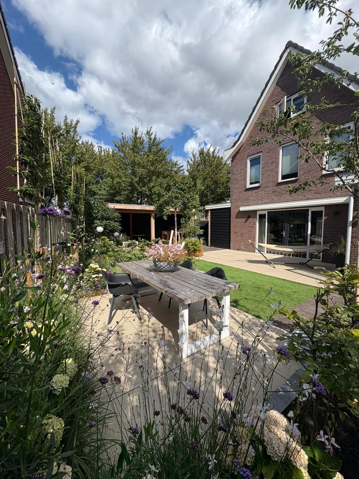 Een achtertuin met een bakstenen huis, een houten tafel, groen en een blauwe hemel met wolken.