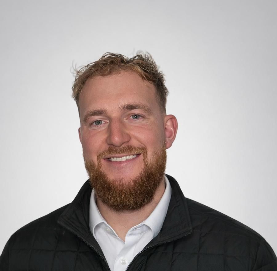 Een man met blond haar, glimlachend, met de armen over elkaar, draagt ​​een zwarte jas met een logo en staat voor een bakstenen muur.