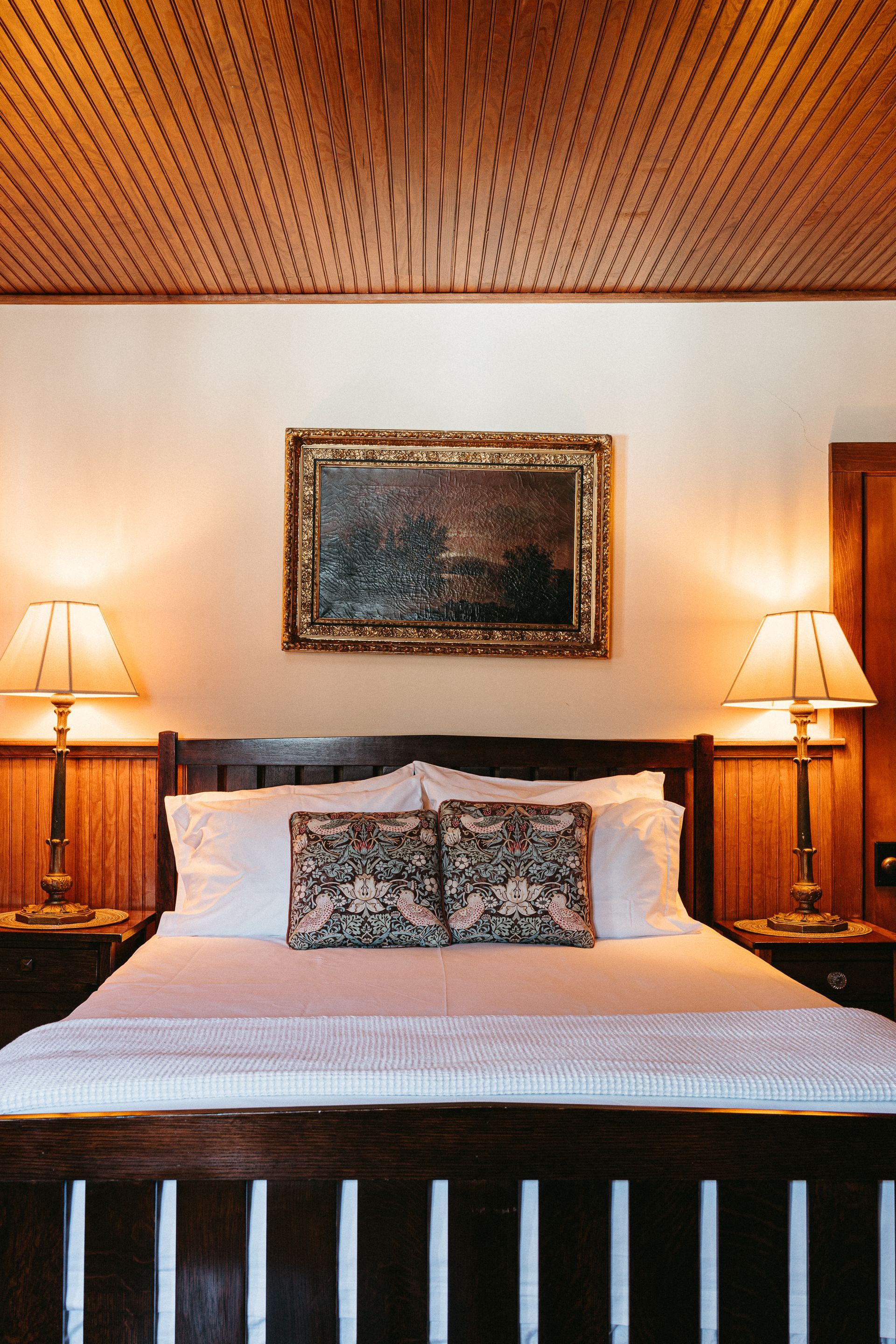 A bedroom with a bed , two lamps , a painting on the wall and a wooden ceiling.