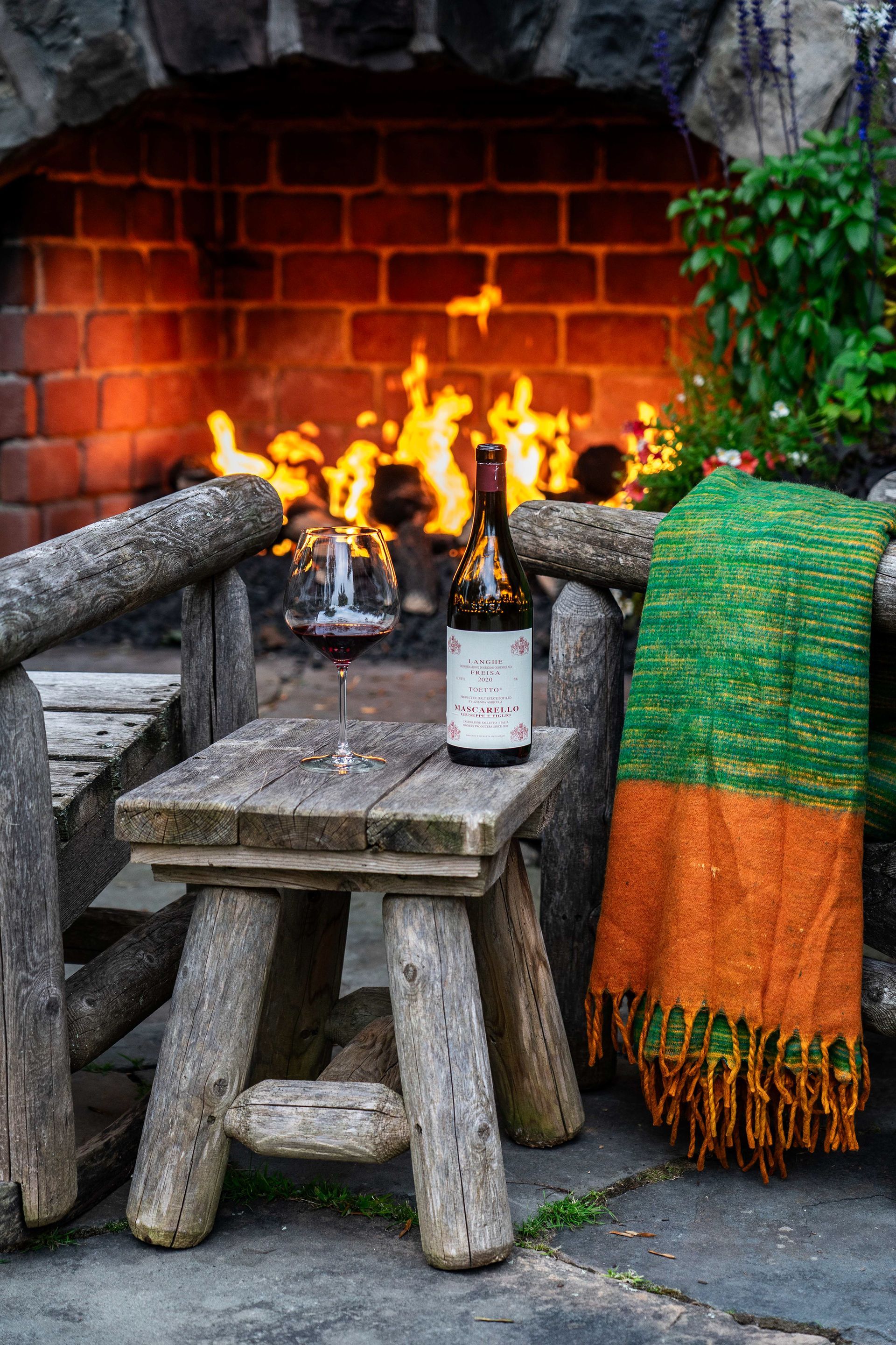 A bottle of wine is sitting on a wooden stool in front of a fireplace.