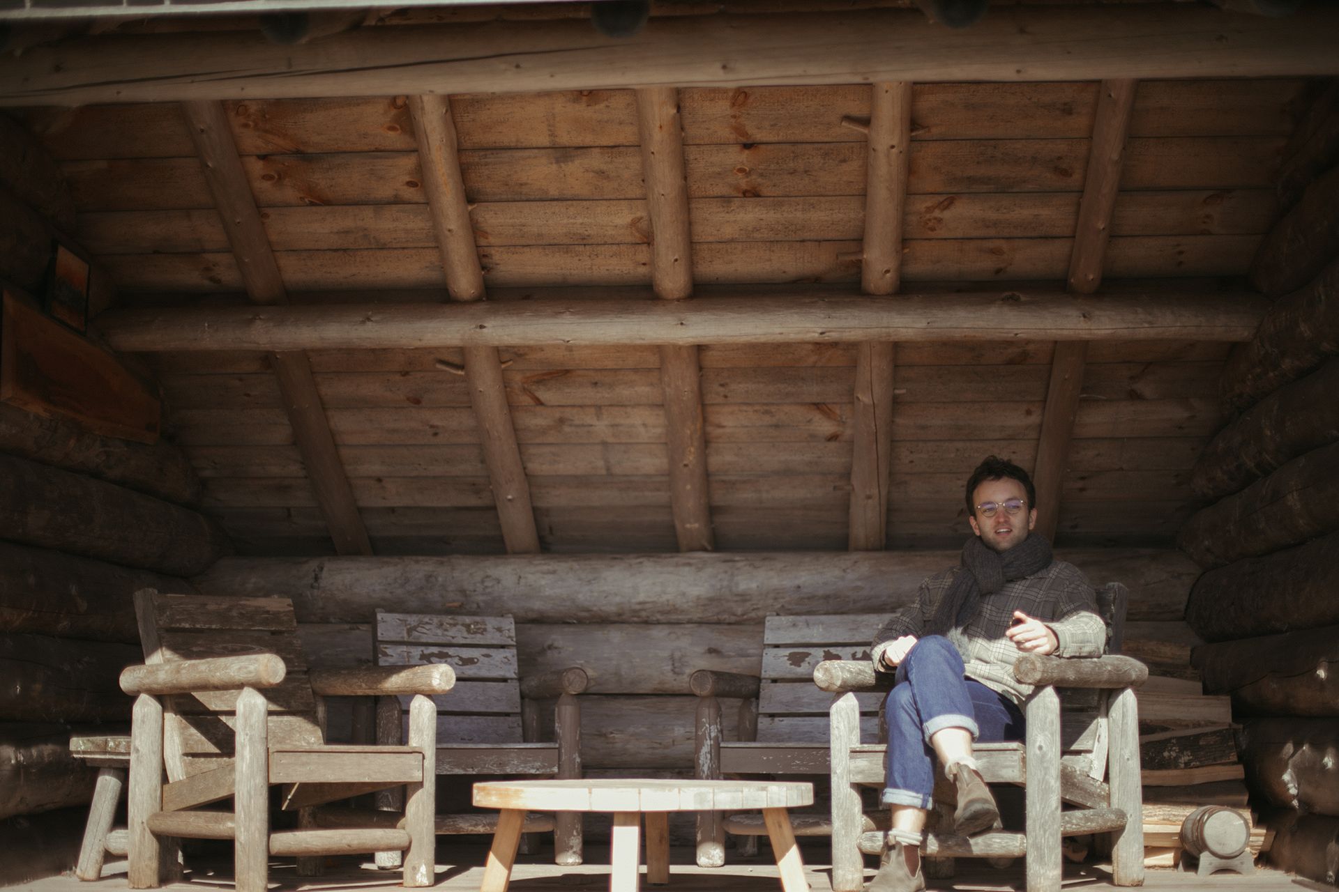 A man is sitting in a wooden chair under a wooden roof.