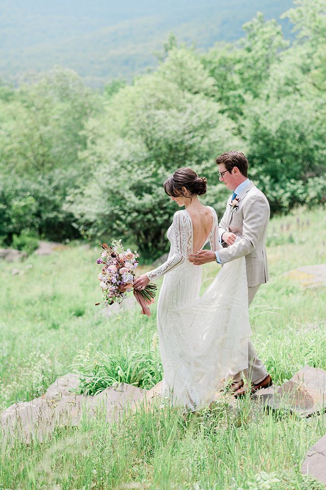 A bride and groom are walking through a grassy field holding hands.
