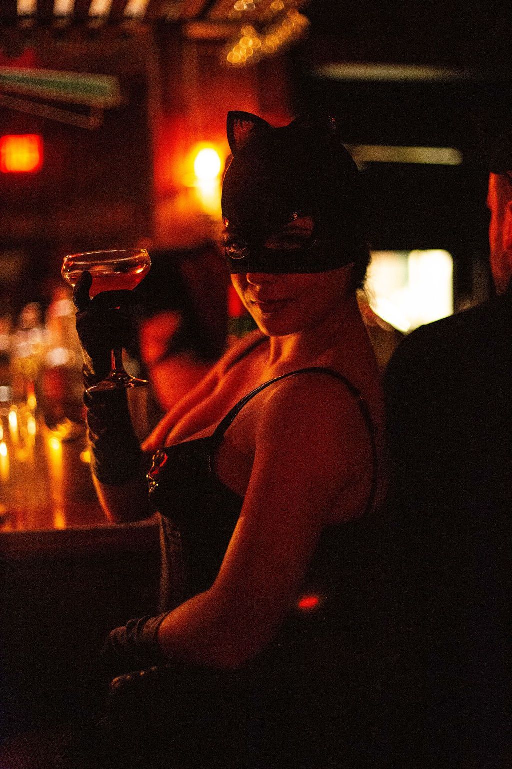 A woman in a cat mask is sitting at a bar holding a drink