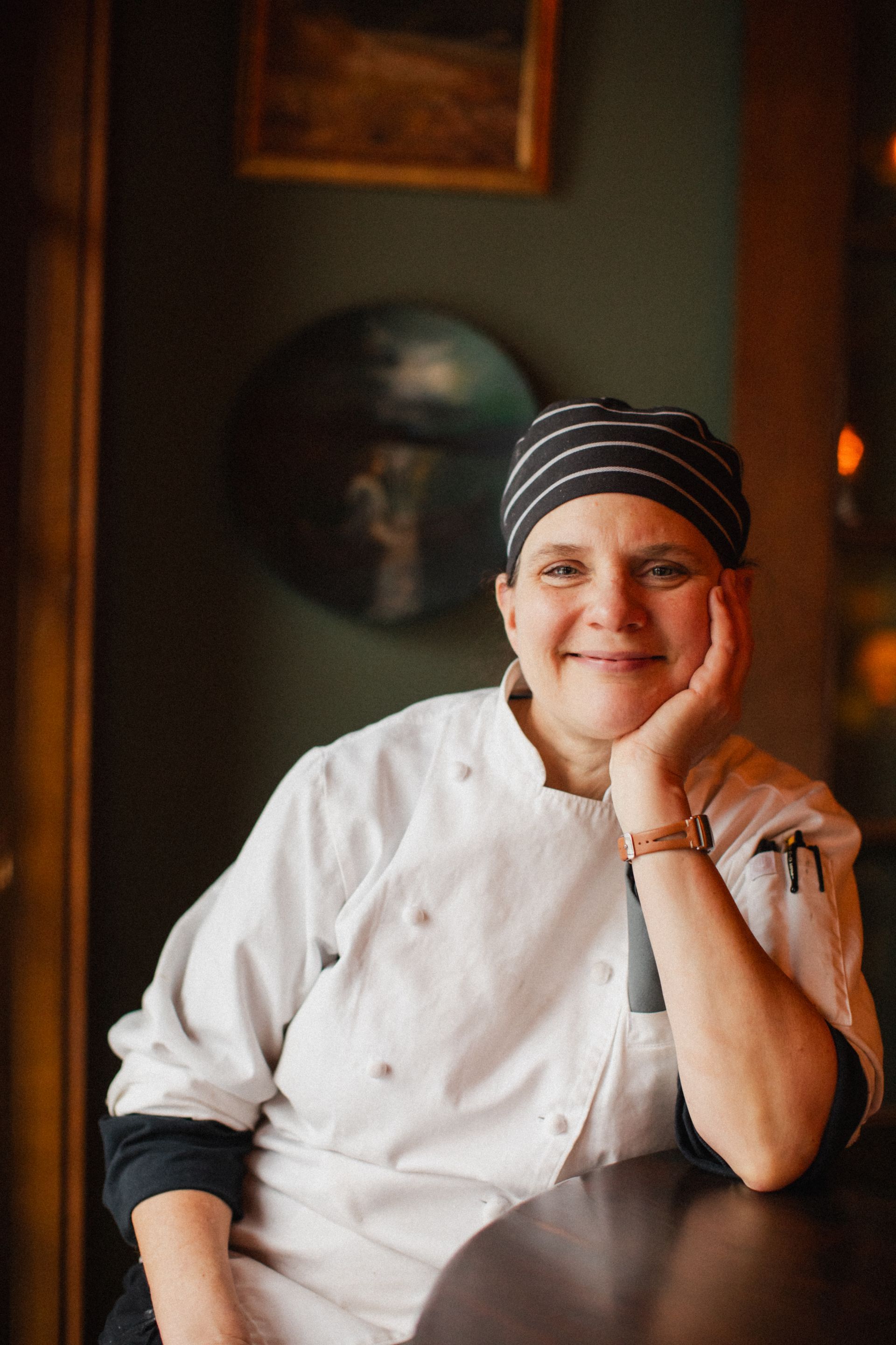 A woman in a chef 's uniform is sitting at a table with her hand on her chin.
