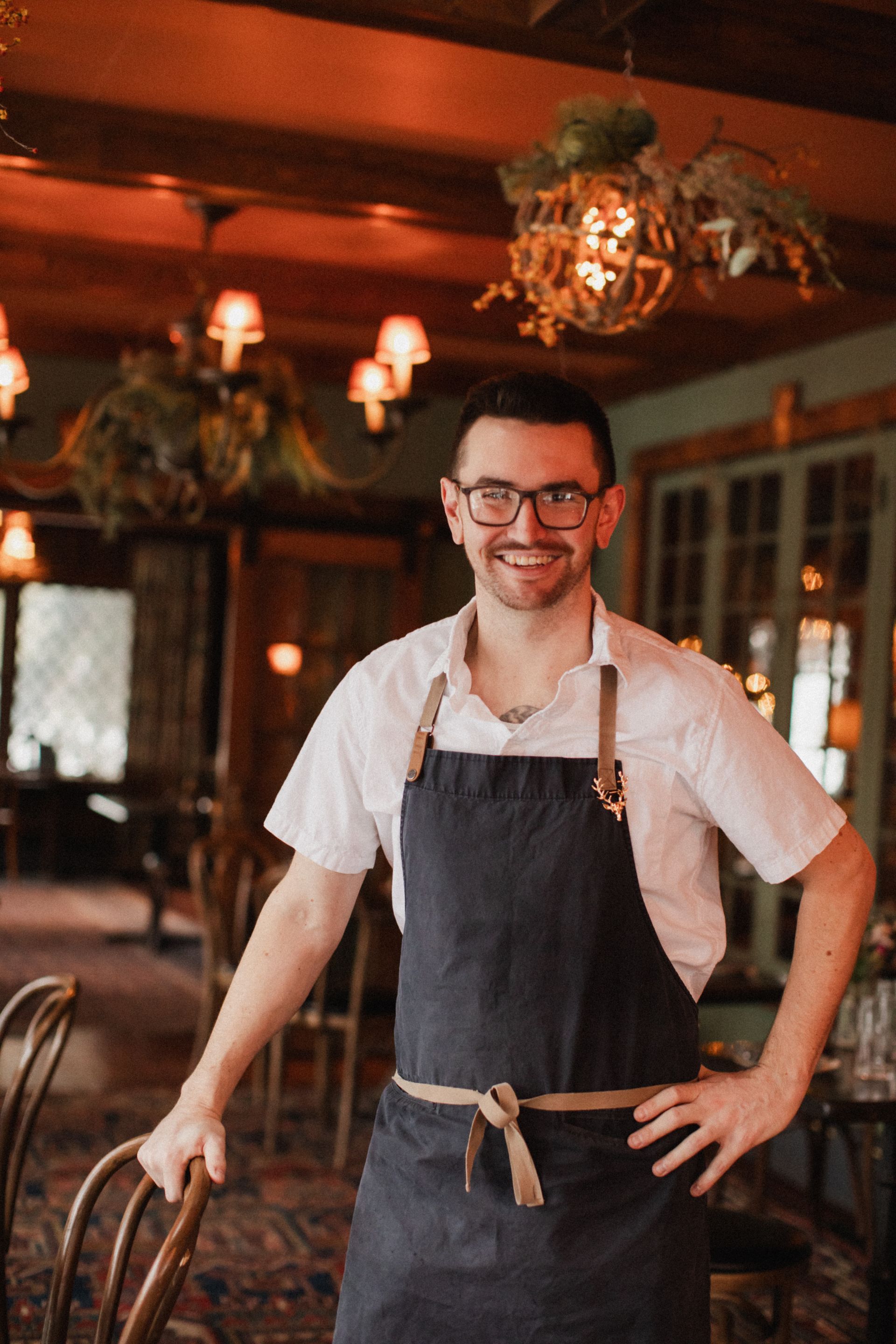 A man wearing an apron and glasses is standing in a restaurant.
