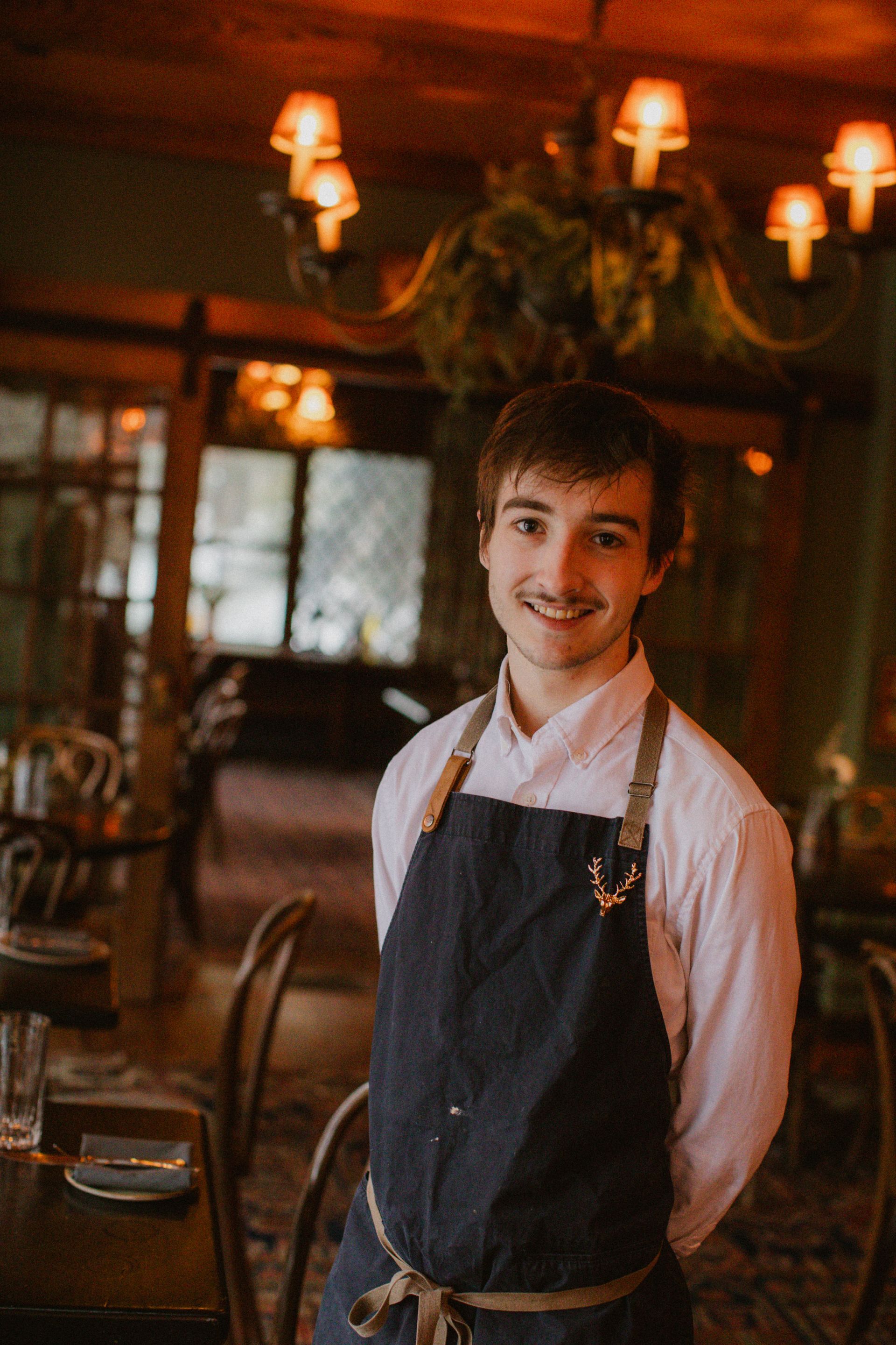 A man in an apron is standing in a restaurant.
