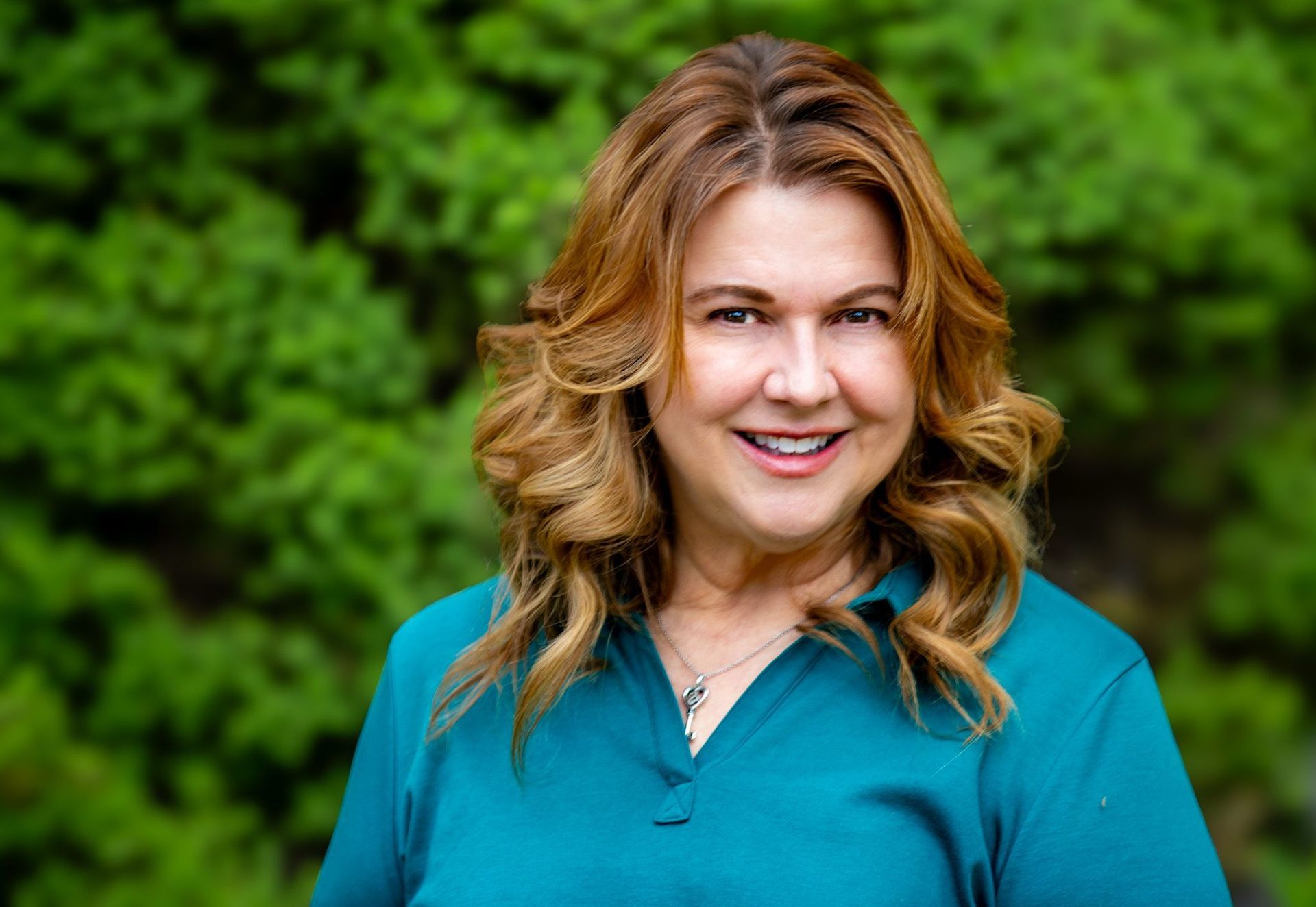 Smiling woman in a teal top standing outdoors against a green leafy background