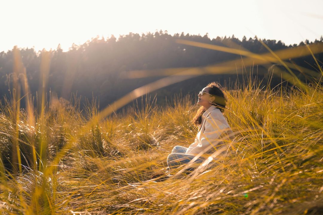 Young woman sitting in tall golden grass at sunset, with sunlit hills and trees in the background. She has dark curly hair and glasses and is wearing light wash jeans and a beige jacket