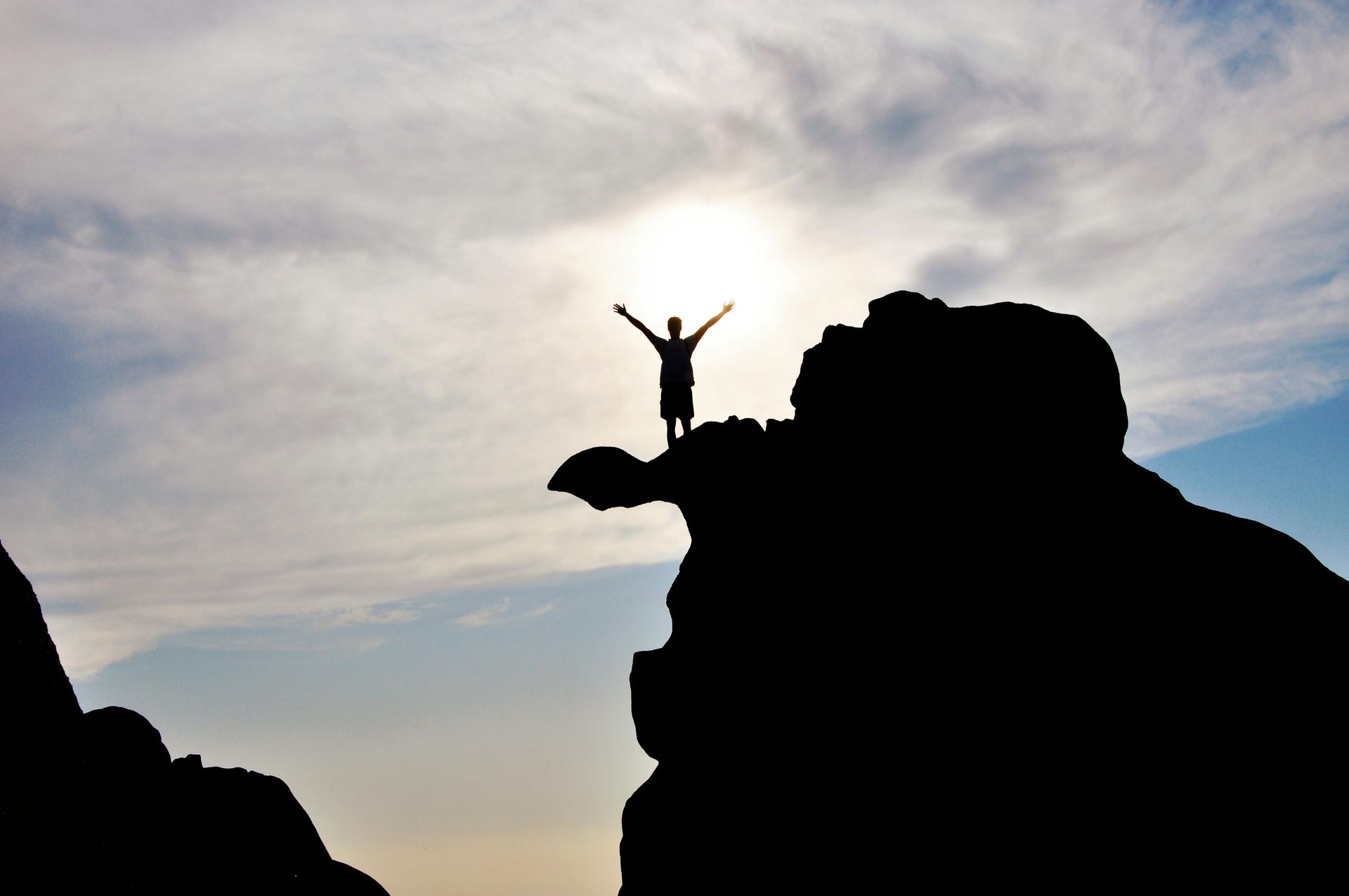 Silhouette of a person standing on rocky cliffs at sunset with arms raised against a cloudy sky