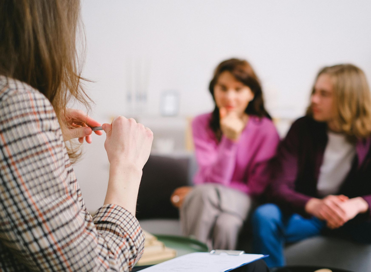 Person speaking to two seated people in a casual meeting room, with notes in the foreground