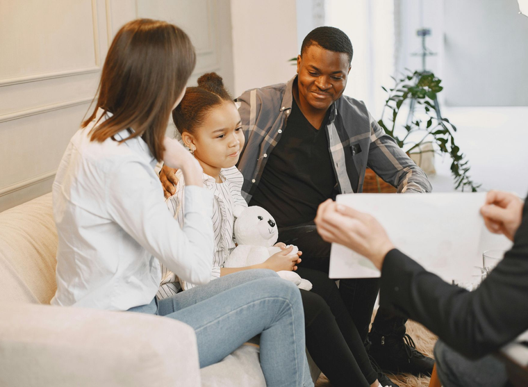 Family talking with a counselor in a bright living room, with a child holding a stuffed animal.