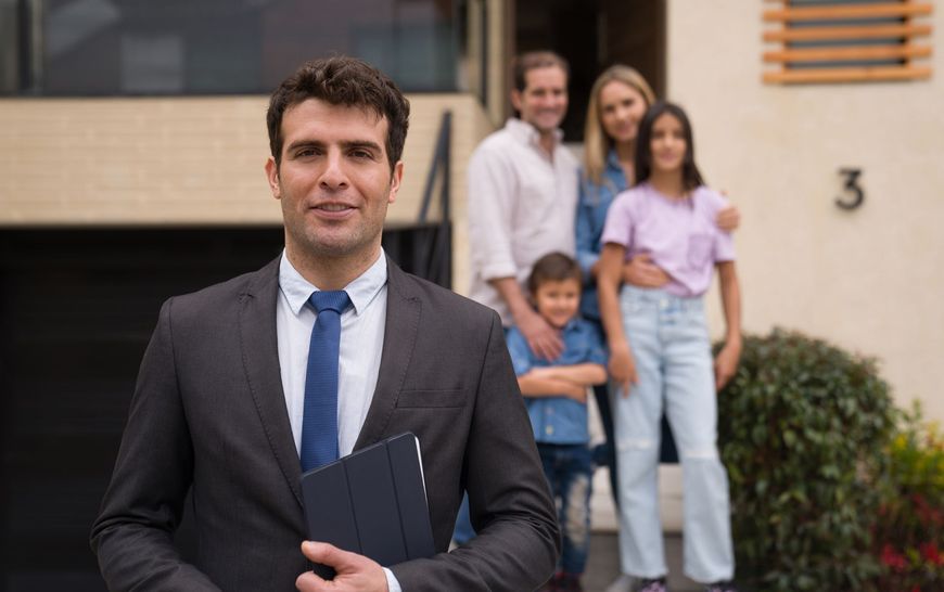 Real estate agent stands in front of a house, a family stands on the porch behind him.