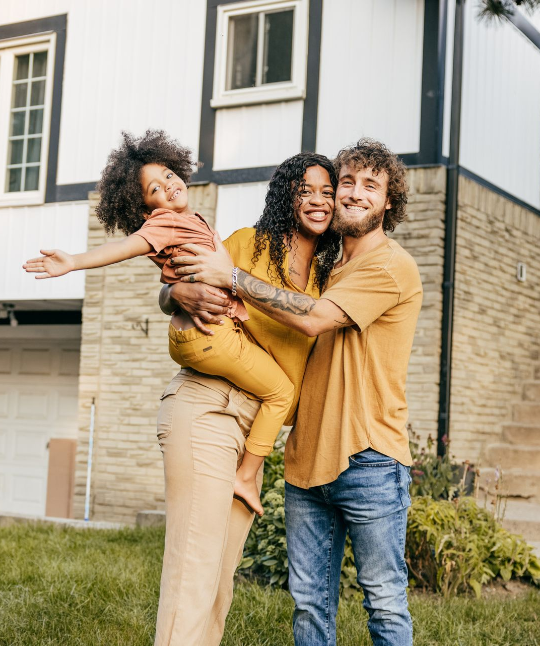 Family of three outside a house, smiling. Woman holding a child, man smiling.