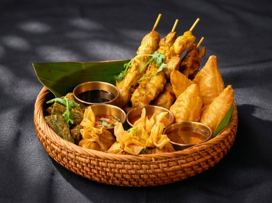 Assorted fried snacks in a woven basket with dipping sauces on a dark background