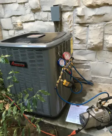Outdoor HVAC unit with gauges and hoses connected, beside a vacuum pump against a stone wall.