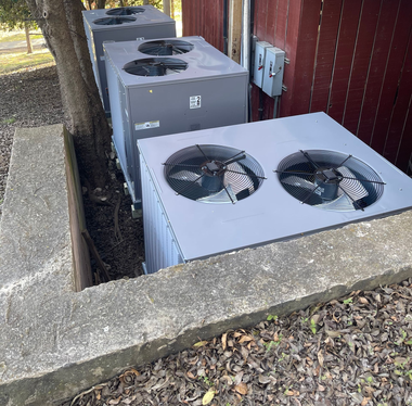 Three outdoor HVAC units beside a red building, viewed over a low concrete wall.