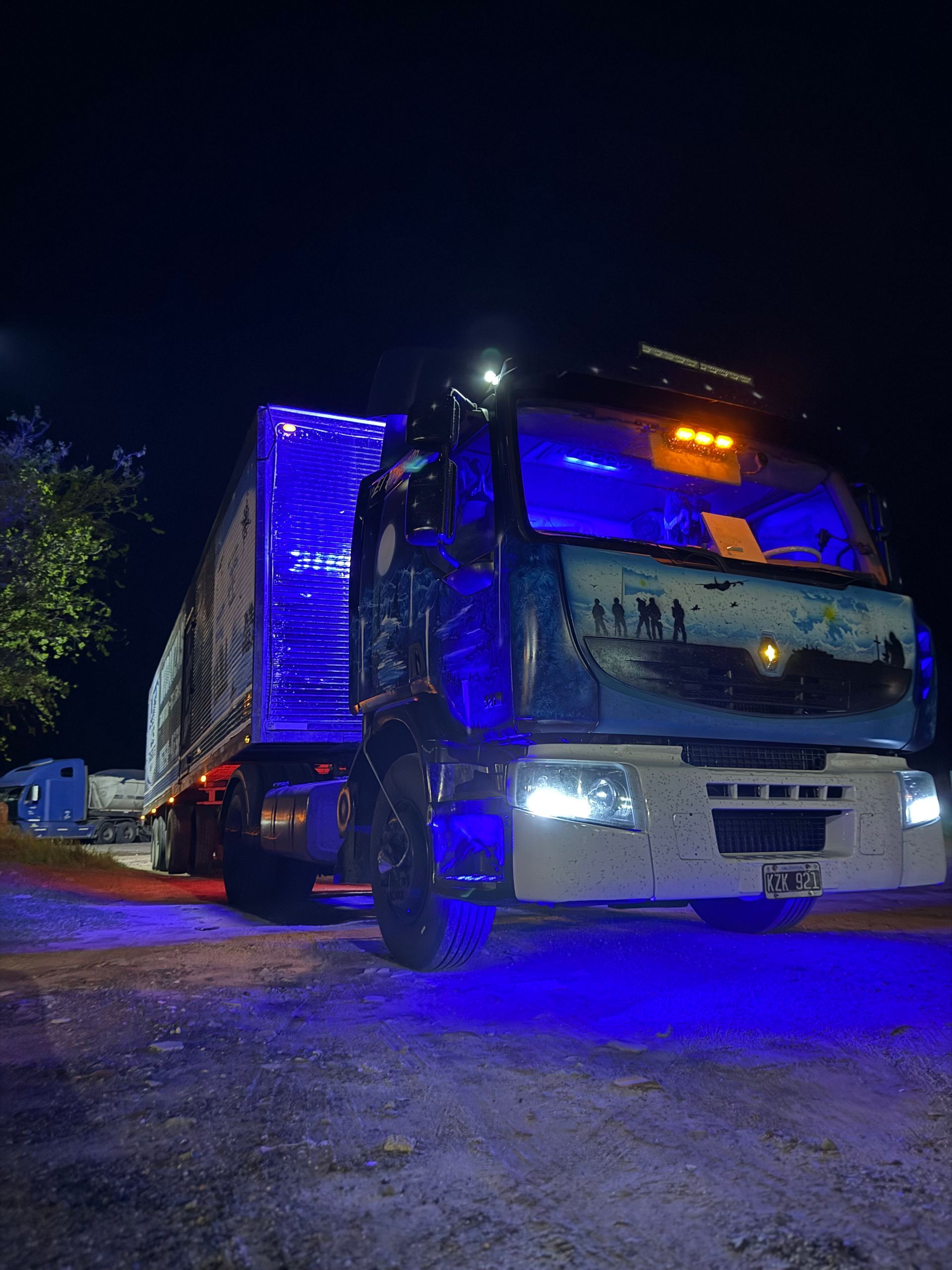 Camión semirremolque iluminado de azul por la noche, con un mural en la cabina, estacionado sobre una superficie oscura.