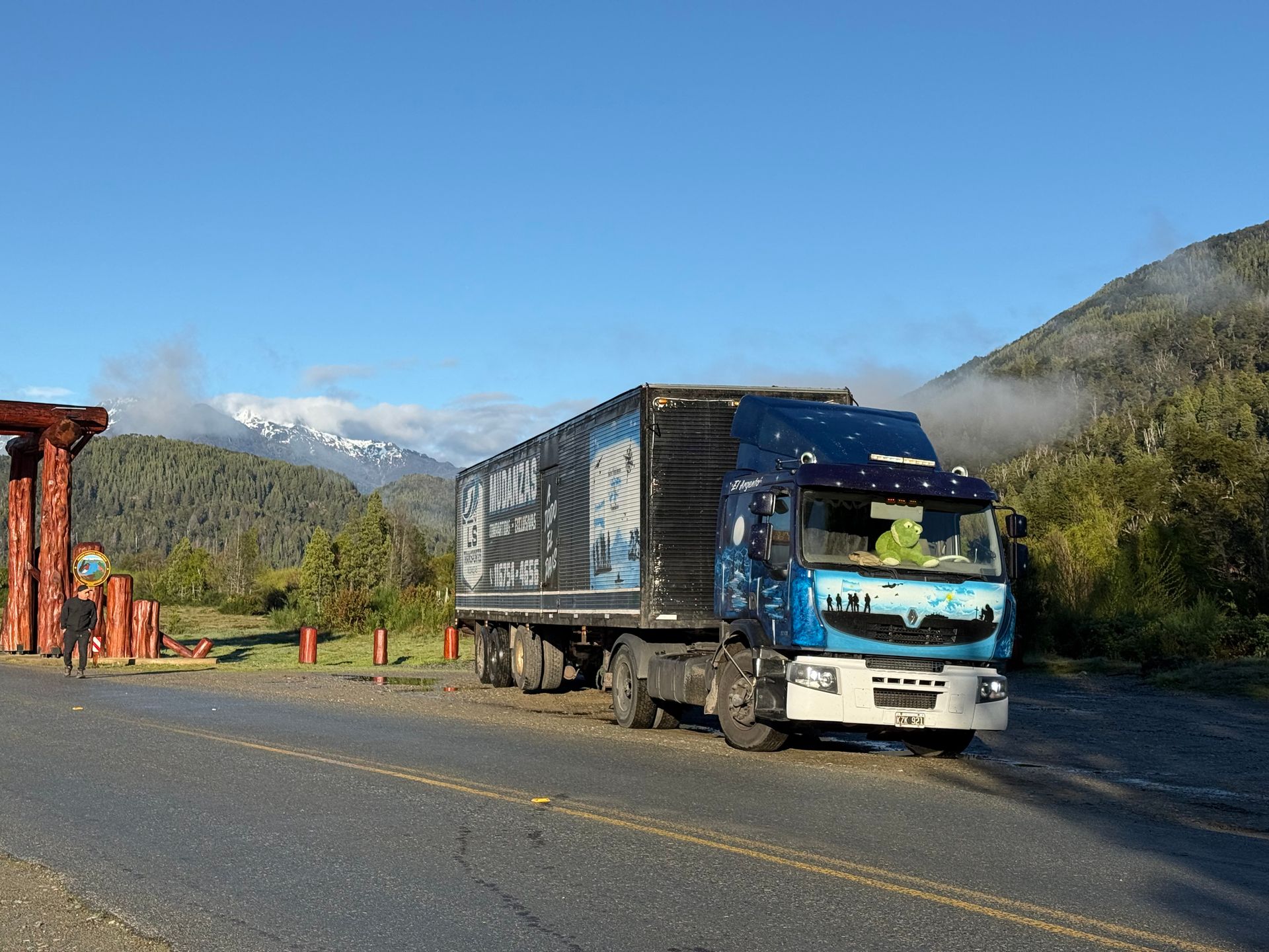 Camión semirremolque estacionado en la carretera cerca de una estructura de madera. Paisaje montañoso y boscoso con nubes al fondo.