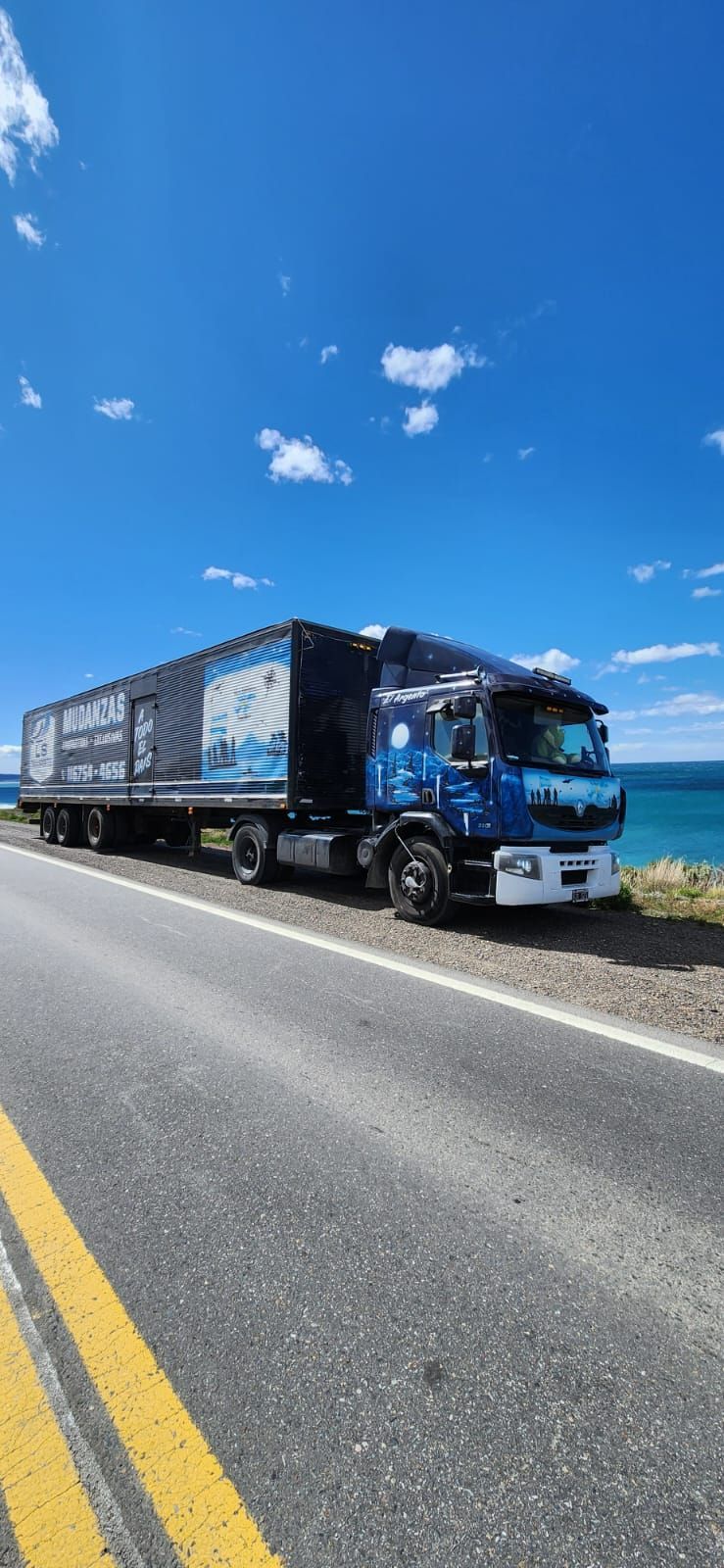 Un camión azul en una carretera junto al océano bajo un cielo azul brillante.