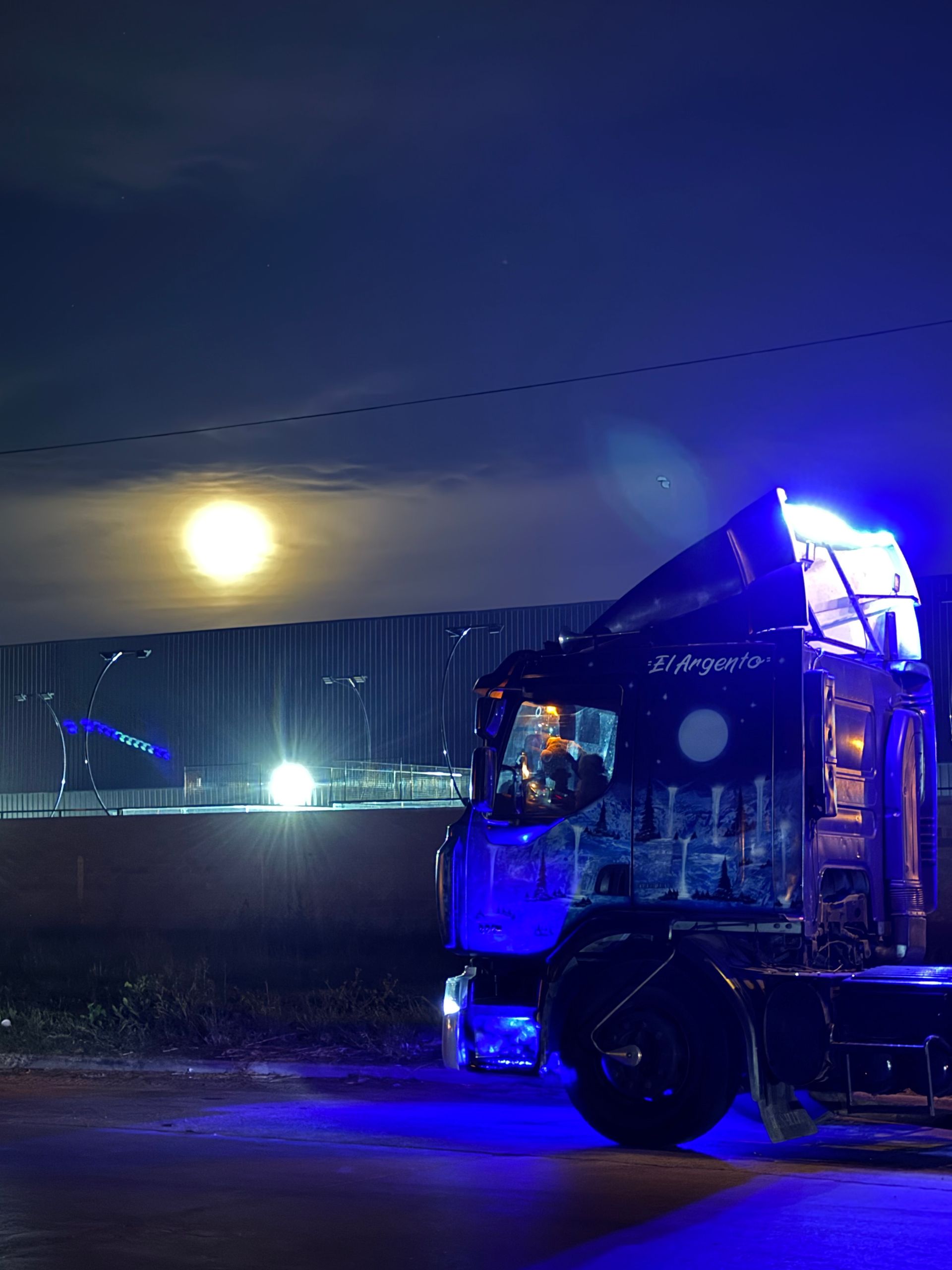 Camión semirremolque iluminado de azul por la noche con la luna llena de fondo.