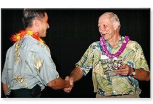 Two men shaking hands with one wearing a lei