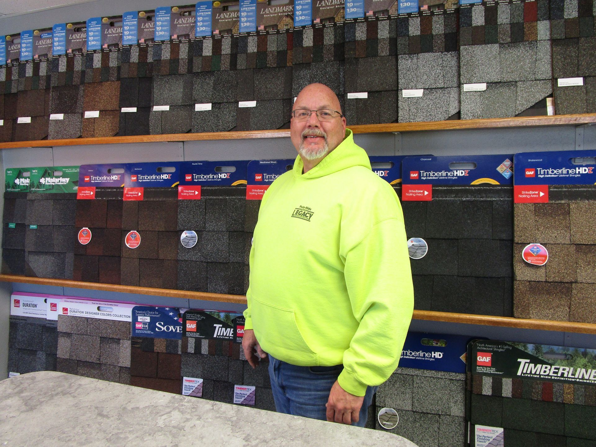 A man in a neon yellow hoodie is standing in front of a display of shingles.