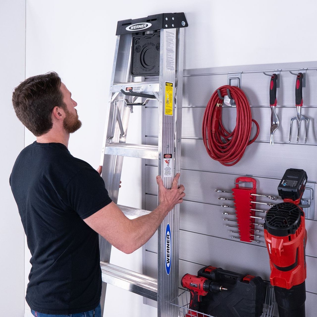 A man is holding a ladder in front of a wall filled with tools.