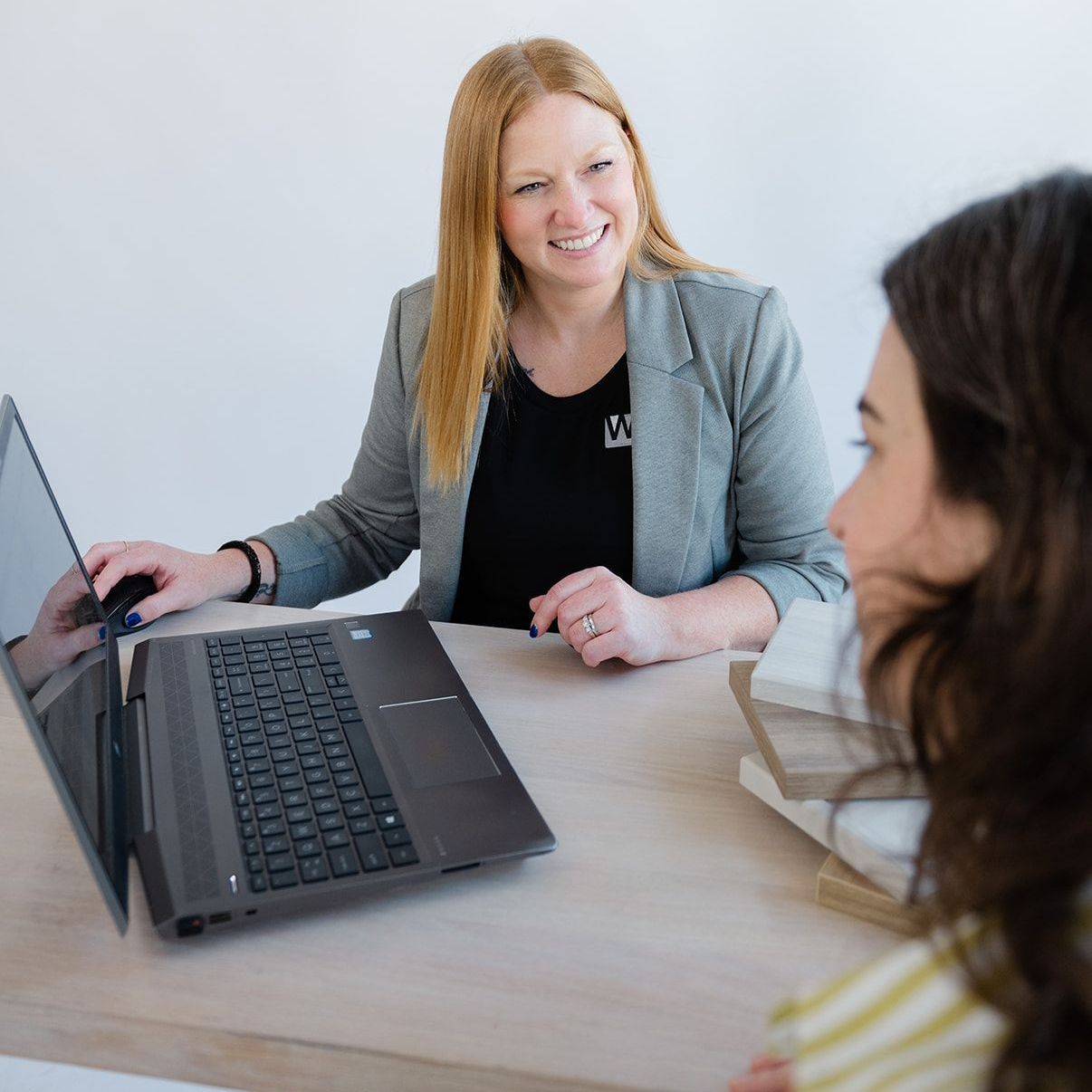 A woman is sitting at a desk with a laptop and talking to another woman.