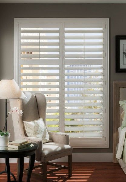 A living room with a chair , table , lamp and window with shutters.