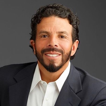 Man with a beard smiles, wearing a suit jacket and white shirt against a gray background.