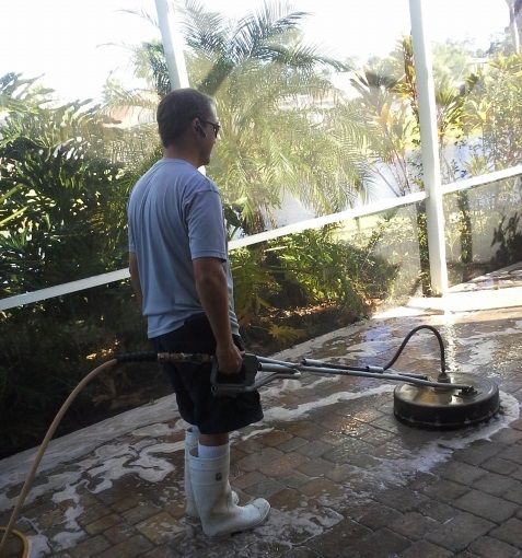 Man pressure washing a brick patio outdoors, wearing boots, water spraying, sunny day.