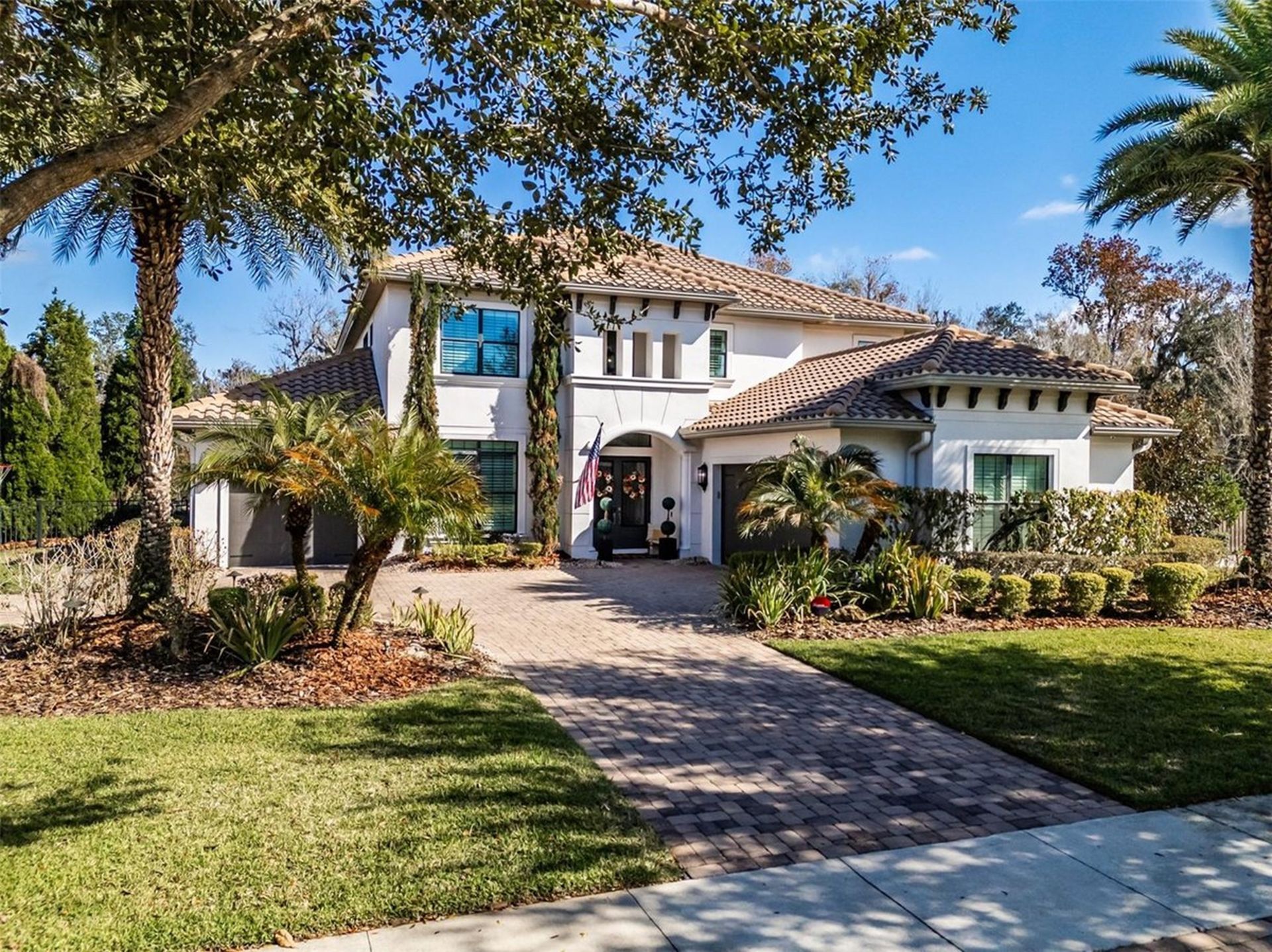 A two-story stucco house with a tiled roof, arched entryway, paved driveway, and palm trees under a bright blue sky.