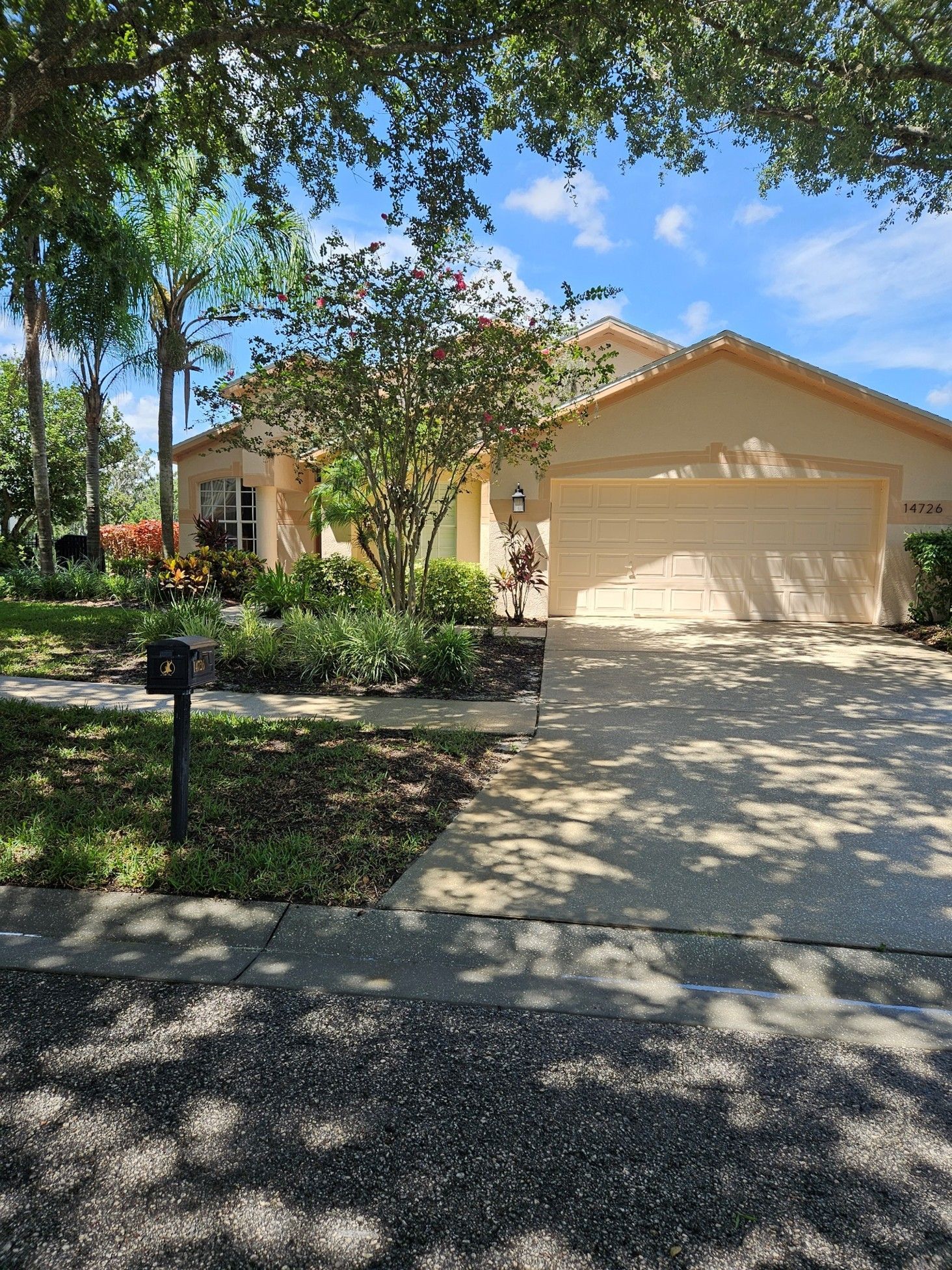 A tan, single-story house with a two-car garage, surrounded by lush trees and landscaping on a sunny day.